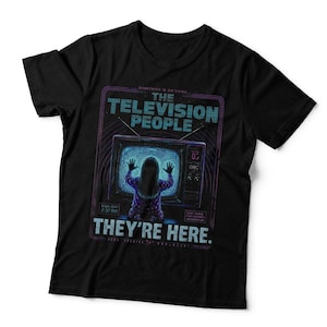 May include: A black t-shirt featuring a graphic of a person facing a television screen with hands pressed against the glass. The text reads THE TELEVISION PEOPLE and THEY'RE HERE, with additional small text and purple line accents.
