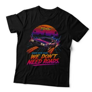 May include: A black t-shirt featuring a graphic of the DeLorean car from Back to the Future, surrounded by lightning and a sunset. The text reads: Where we're going, we don't need roads. The design uses vibrant orange, purple, and blue colours.