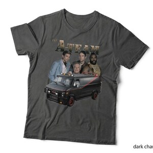 May include: A dark charcoal grey short-sleeve t-shirt featuring a graphic print of the four main characters from The A-Team positioned above their iconic black van with a red stripe. The text The A-Team is printed in a metallic gold font above.