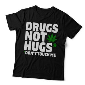 May include: A black short-sleeved t-shirt featuring the white text DRUGS NOT HUGS with a green cannabis leaf icon and a small green asterisk. Below this, the text DON'T TOUCH ME is printed in white, accompanied by a small green asterisk.