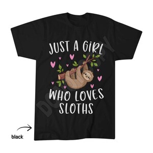 May include: A black short-sleeved t-shirt featuring a graphic of a brown sloth hanging from a green branch. The text reads JUST A GIRL WHO LOVES SLOTHS in white capital letters, surrounded by small pink hearts scattered across the front design.