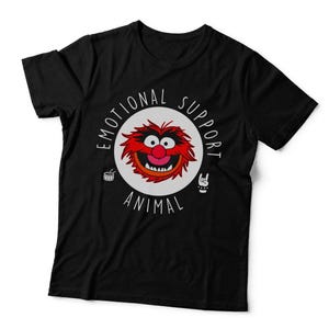 May include: A black short-sleeved t-shirt featuring a circular graphic with a red, furry character face in the centre. The text "EMOTIONAL SUPPORT ANIMAL" arches around the circle, accompanied by a small drum icon and a hand making a rock sign.