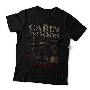 May include: A black short-sleeved t-shirt featuring a graphic print with the text THE CABIN IN THE WOODS at the top and YOU'VE GOT TO BE KIDDING ME at the bottom. The design includes a grid of various horror-themed character illustrations.
