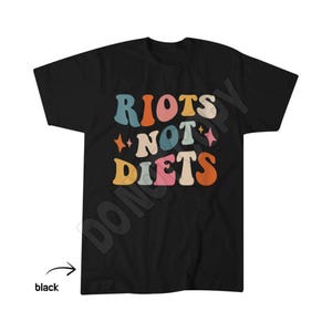 May include: A black short-sleeved t-shirt featuring the text RIOTS NOT DIETS in a multi-coloured, wavy, retro-style font. The design includes small four-pointed star icons interspersed between the words in shades of blue, orange, pink, and cream.