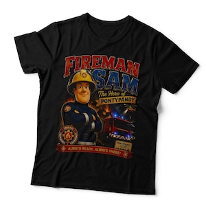 May include: A black short-sleeved t-shirt featuring a graphic of Fireman Sam wearing a yellow helmet. The text reads Fireman Sam The Hero of Pontypandy, Always Ready, Always There! A red fire engine with blue lights is visible in the background.