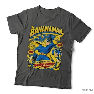 May include: A dark charcoal grey t-shirt featuring a colourful graphic of the comic book character Bananaman. The design includes the text BANANAMAN and POWERED BY PURE BANANA ENERGY, along with illustrations of the hero, bananas, and comic sound effects.
