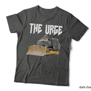 May include: A dark charcoal grey short-sleeve t-shirt featuring the white text THE URGE printed above a graphic of a yellow and black bulldozer. The shirt has a crew neckline and a standard fit, showcasing a construction vehicle design.