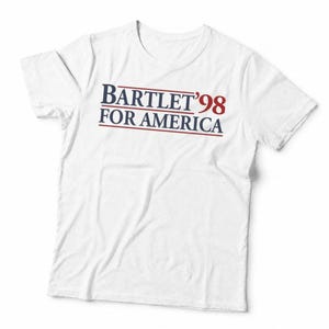 May include: A white short-sleeved t-shirt featuring the text BARTLET '98 FOR AMERICA printed in dark blue and red. The design includes horizontal lines above and below the text, creating a clean, centred graphic on the chest of the garment.