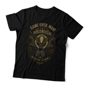 May include: A black short-sleeved t-shirt featuring a graphic print with the text "GAME OVER, MAN!" and "COLONIAL MARINES". The design includes a central alien creature head, two crossed rifles, and two dog tags, all rendered in muted yellow and olive tones.