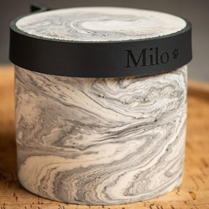 Puede incluir: Un recipiente cilíndrico con un patrón de mármol blanco y gris. La tapa está asegurada por una banda negra que presenta el nombre Milo en relieve y un pequeño icono de huella. El recipiente tiene una apariencia lisa, similar a la piedra, y una parte superior plana.