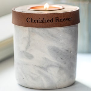 May include: A white marble candle holder with a brown leather band that says "Cherished Forever". The candle holder has a small, round opening for a tea light candle.