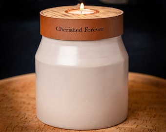 May include: A light beige ceramic jar with a smooth finish, topped with a circular wooden lid featuring a central tea light candle holder. A brown leather band wraps around the top, embossed with the black text Cherished Forever.