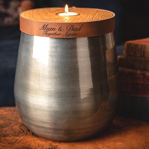 May include: A metallic silver urn with a brushed finish, topped with a circular wooden lid featuring a central tea light candle. A brown leather band wraps around the lid, engraved with the text Mum & Dad Together Again.