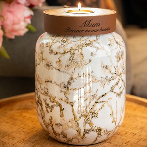Op de afbeelding: Een witte keramische urn met een bloemenpatroon en een houten deksel. De deksel heeft een bruine band met de tekst "Mum Forever in our hearts". Bovenop staat een brandende kaars. Roze bloemen zijn op de achtergrond.