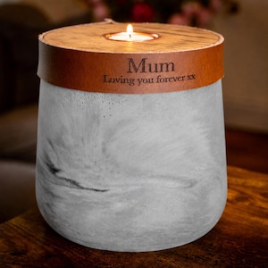 Puede incluir: Un portavelas cilíndrico de hormigón con un acabado gris jaspeado y una banda de cuero marrón envuelta en la parte superior. El cuero presenta el texto grabado Mum y Loving you forever xx. Una vela de té encendida se sitúa en el centro.