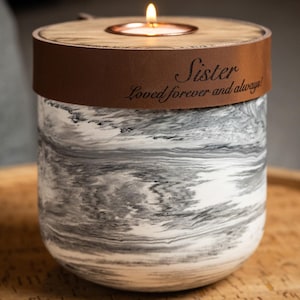 Puede incluir: Un portavelas cilíndrico con un patrón de mármol blanco y gris. Una banda de cuero marrón envuelve la parte superior, con el texto grabado "Sister Loved forever and always". Una vela de té encendida se sitúa en la tapa de madera central.