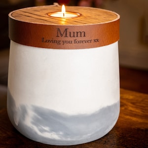 Puede incluir: Un portavelas de hormigón blanco y gris con una banda de cuero marrón alrededor de la parte superior. El cuero tiene grabado el texto Mum y Loving you forever xx. Una vela de té encendida se sitúa en el centro de la tapa de madera.