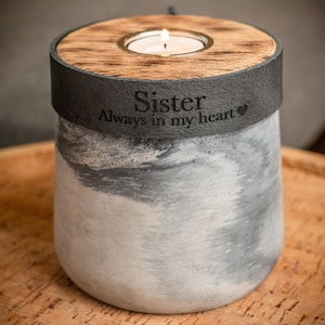 Puede incluir: Un portavelas conmemorativo de hormigón gris y blanco con una tapa de madera y una pequeña vela de té. El portavelas tiene una banda de cuero negro con la inscripción "Hermana Siempre en mi corazón" y un pequeño símbolo de corazón.