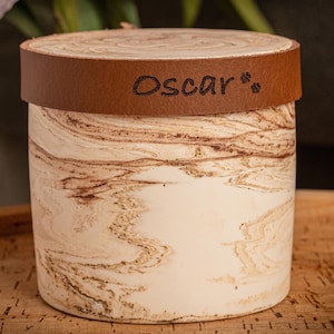 Puede incluir: Un recipiente cilíndrico con un acabado jaspeado en crema y marrón, que presenta una banda de cuero marrón alrededor de la tapa. La banda de cuero está grabada con el nombre Oscar y dos pequeños iconos de huellas en un color oscuro.