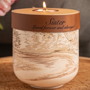 Puede incluir: Un portavelas cilíndrico con un patrón de mármol blanco y marrón. Una banda de color cobre rodea la parte superior, con el texto grabado "Sister" y "Loved forever and always!". Una vela de té encendida se encuentra en la tapa de madera.