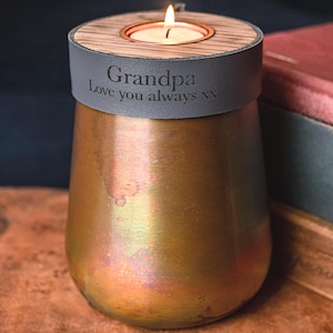 Puede incluir: Un portavelas de tono cobre con cuerpo cónico y una banda de cuero gris oscuro con el texto grabado "Grandpa Love you always xx". La parte superior tiene un inserto de madera con una vela de té encendida en el centro.