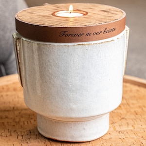 May include: White ceramic urn with a wooden lid and a leather band that says "Forever in our hearts". The lid has a small candle holder with a lit candle.