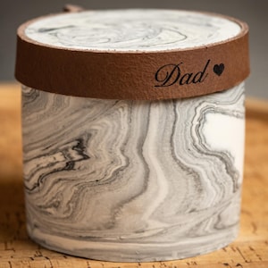 Puede incluir: Un recipiente cilíndrico con un patrón de mármol blanco y gris. La tapa está asegurada por una banda de cuero marrón con el texto grabado "Dad" seguido de un pequeño símbolo de corazón. El recipiente tiene un acabado liso y pulido.