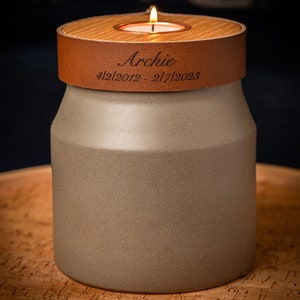 May include: A cylindrical beige ceramic vessel topped with a brown leather band featuring the engraved name Archie and the dates 4/2/2012 - 2/7/2023. A small lit tea light candle sits in the center of the wooden lid covering the top opening.