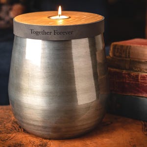 Könnte beinhalten: Eine silberfarbene Metallurne mit konischem Körper und Holzdeckel. Der Deckel verfügt über einen zentralen Teelichthalter mit brennender Kerze. Ein dunkelgraues Band am oberen Rand der Urne ist mit dem Text Together Forever in Serifenschrift graviert.