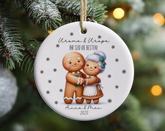 May include: A white ceramic Christmas ornament with a gingerbread couple holding hands. The ornament has the text "Uroma & Uropa. IHR SEID DIE BESTEN!" and "Anna & Max 2023".