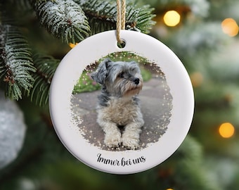 May include: White ceramic Christmas ornament with a photo of a small, gray and brown dog with a sparkly background. The text "Immer bei uns" is printed on the ornament.