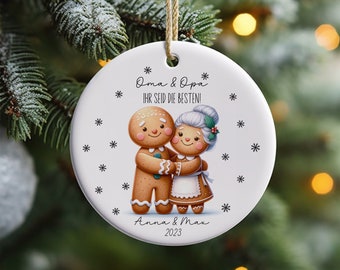 May include: A white ceramic Christmas ornament with a gingerbread couple holding hands. The ornament has the text "Oma & Opa IHR SEID DIE BESTEN!" and "Anna & Max 2023".