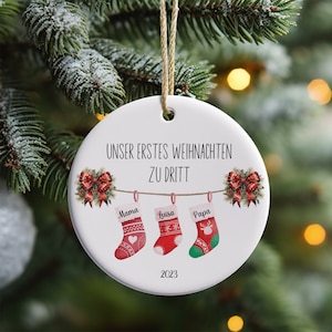 May include: White ceramic Christmas ornament with the text "Unser erstes Weihnachten zu dritt" (Our first Christmas as a family of three) and the year 2023. Three red and green stockings with the names "Mama", "Luisa", and "Papa" are hanging from a string.