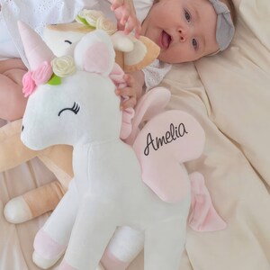 Pu&ograve; includere: Un giocattolo di peluche a forma di unicorno bianco con zoccoli rosa, una criniera rosa e un'ala a forma di cuore rosa con il nome "Amelia" ricamato. L'unicorno &egrave; sdraiato su una coperta color crema.