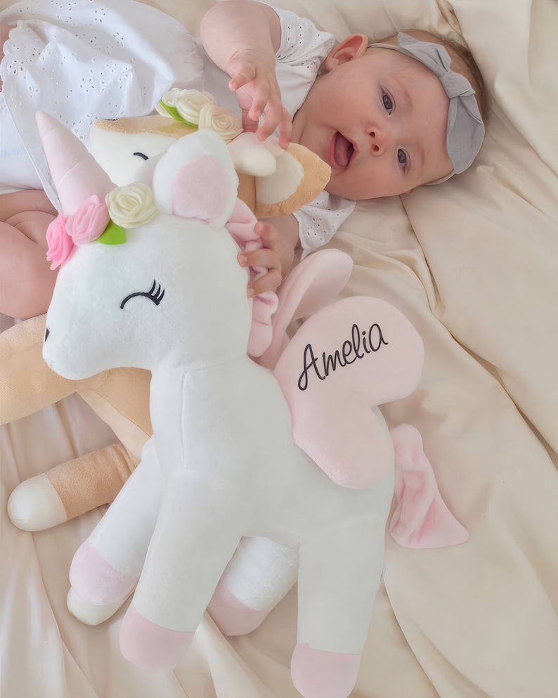 Pu&ograve; includere: Un giocattolo di peluche a forma di unicorno bianco con zoccoli rosa, una criniera rosa e un'ala a forma di cuore rosa con il nome "Amelia" ricamato. L'unicorno &egrave; sdraiato su una coperta color crema.