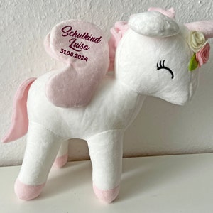 Pu&ograve; includere: Unicorno di peluche bianco con ali rosa e coda rosa. L'unicorno indossa una corona di fiori rosa e un testo ricamato che dice "Schulkind Luisa 31.08.2024".