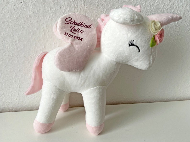 Pu&ograve; includere: Unicorno di peluche bianco con ali rosa e coda rosa. L'unicorno indossa una corona di fiori rosa e un testo ricamato che dice "Schulkind Luisa 31.08.2024".