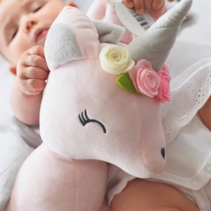 Pu&ograve; includere: Un giocattolo di peluche unicorno rosa con un corno grigio e una corona di fiori. La corona ha fiori bianchi, rosa e verdi.