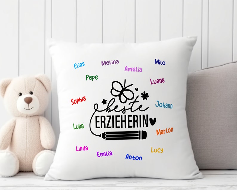 May include: White pillow with black text that says "*beste ERZIEHERIN*" with a pencil and a butterfly design. The pillow is surrounded by names written in different colors.