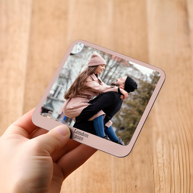 May include: A square coaster featuring a photograph of a couple in London. The coaster has a pale pink border and the text "London 2025". The photo shows a woman in a pink coat and hat being carried by a man in a black coat and hat.