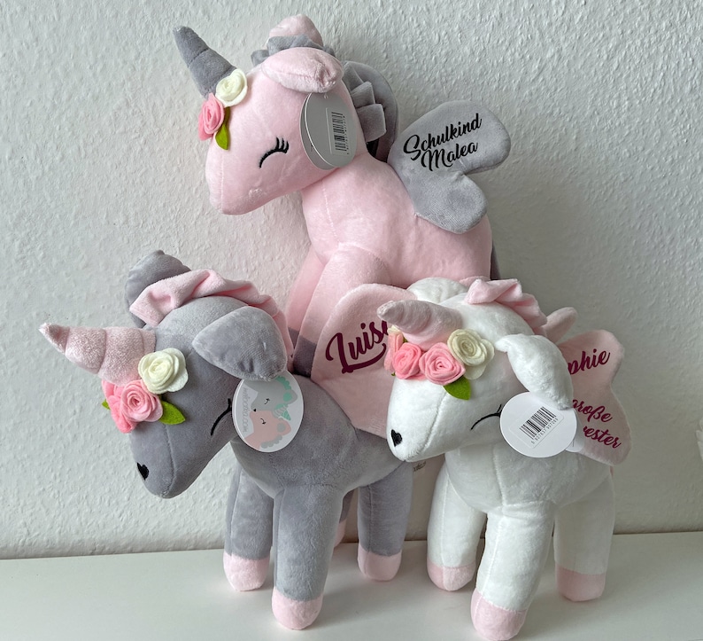 Pu&ograve; includere: Tre giocattoli di peluche a forma di unicorno con nomi ricamati. L'unicorno rosa ha un'ala grigia e il testo "Schulkind Malea". L'unicorno grigio ha il testo "Luis". L'unicorno bianco ha il testo "Sophie gro&szlig;e erster".