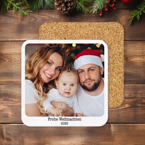May include: A square coaster with a family photo, featuring a smiling woman, a man in a Santa hat, and a baby. The coaster has the text "Frohe Weihnachten 2025" and sits on a cork base, with a wooden background.