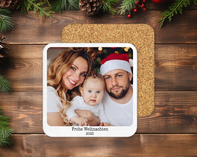 May include: A square coaster with a family photo, featuring a smiling woman, a man in a Santa hat, and a baby. The coaster has the text "Frohe Weihnachten 2025" and sits on a cork base, with a wooden background.