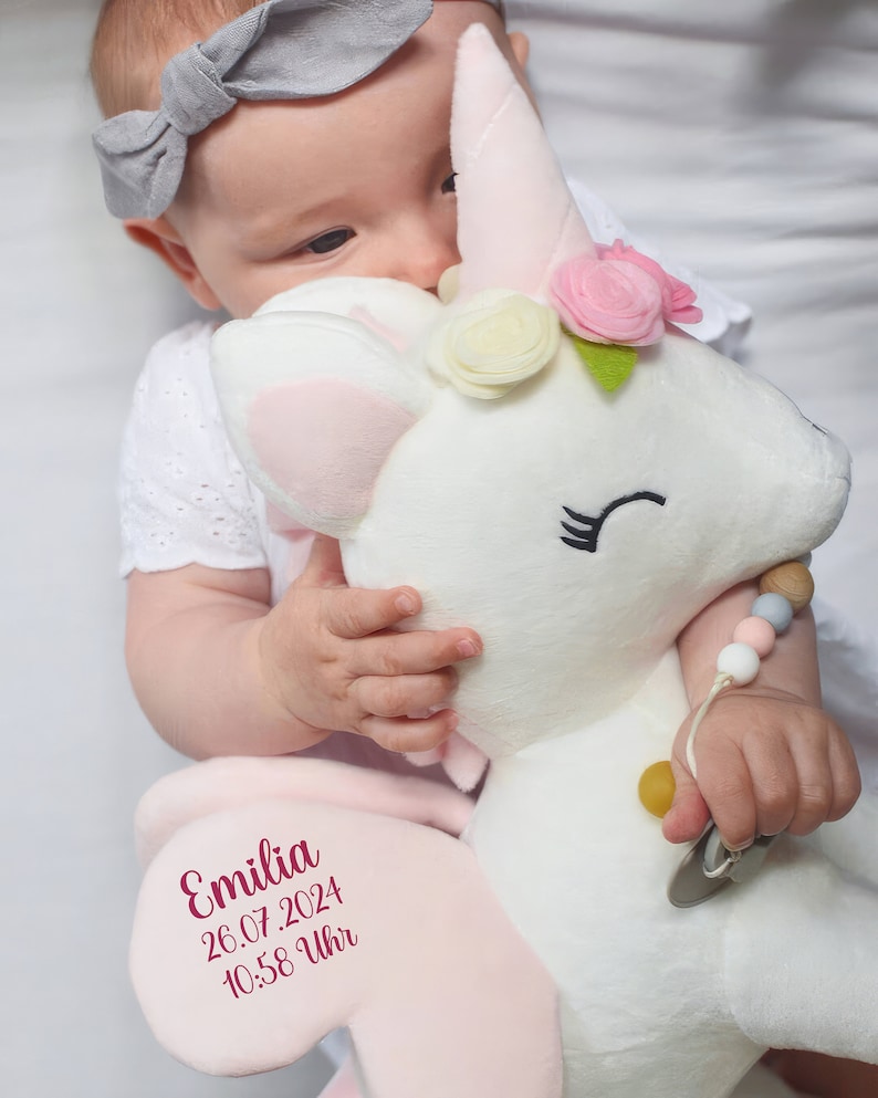 Pu&ograve; includere: Un giocattolo di peluche a forma di unicorno bianco con orecchie rosa, un corno rosa e una corona di fiori. L'unicorno ha una toppa rosa sul fianco con il testo "Emilia 26.07.2024 10:58 Uhr". Un bambino tiene l'unicorno.