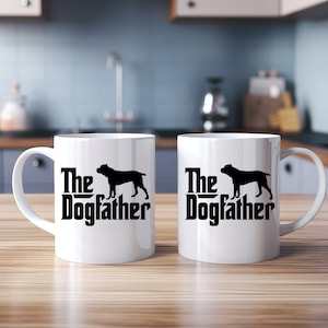 Könnte beinhalten: Zwei weiße Keramikbecher mit schwarzen Henkeln. Die Becher haben eine schwarze Silhouette eines Hundes und den Text "The Dogfather" aufgedruckt.