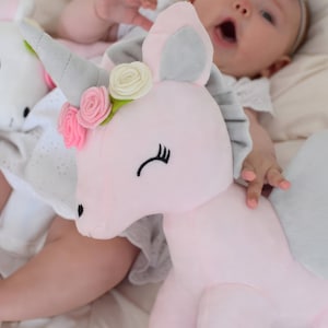 Pu&ograve; includere: Un giocattolo di peluche a forma di unicorno rosa con un corno grigio, orecchie grigie e una corona di fiori bianchi. L'unicorno &egrave; sdraiato su una superficie bianca con un bambino che lo tiene.