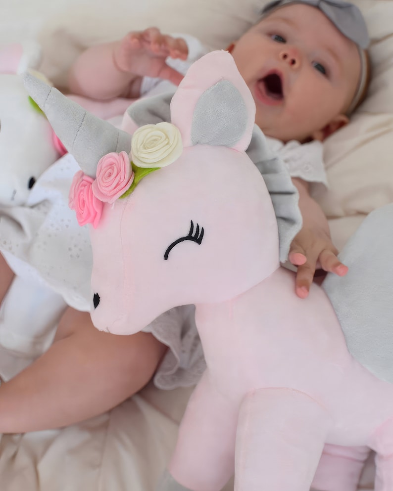 Pu&ograve; includere: Un giocattolo di peluche a forma di unicorno rosa con un corno grigio, orecchie grigie e una corona di fiori bianchi. L'unicorno &egrave; sdraiato su una superficie bianca con un bambino che lo tiene.