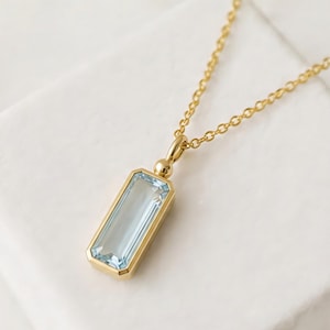 May include: A gold necklace featuring a rectangular, pale blue gemstone pendant. The pendant is set in a gold bezel and suspended from a fine gold chain. The necklace is presented on a white surface.