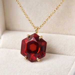 Peut inclure: Un collier à chaîne dorée avec un grand pendentif rouge hexagonal facetté. Le pendentif est serti de griffes dorées et présenté dans un écrin blanc avec un intérieur crème.