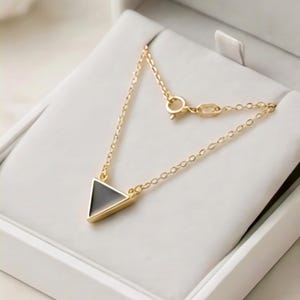 May include: A gold necklace featuring a triangular pendant with a black centre and gold edging. The necklace is presented in a white jewellery box.